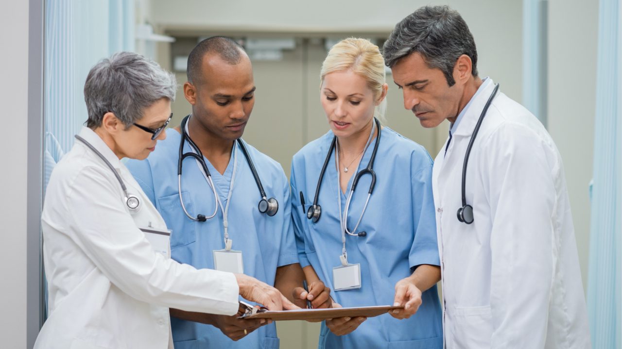 Four healthcare professionals with stethoscopes looking at clipboard as one points.