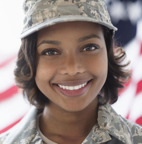 A service member wearing camouflage with an American flag in the background.