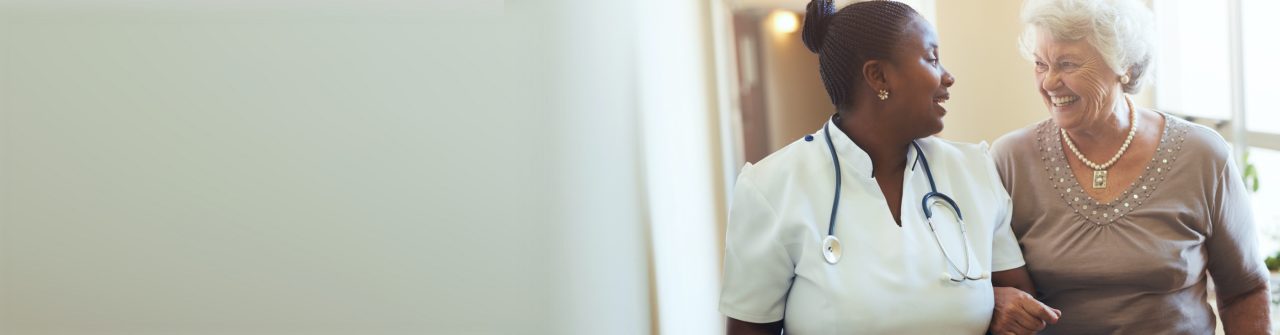 Nurse wearing a white uniform with a stethoscope around her neck walking down a hallway with a smiling patient.