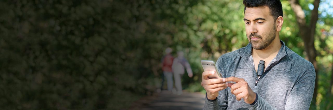 Man wearing BodyGuardian MINI PLUS Remote Cardiac Monitor as he looks at his mobile phone in a park.
