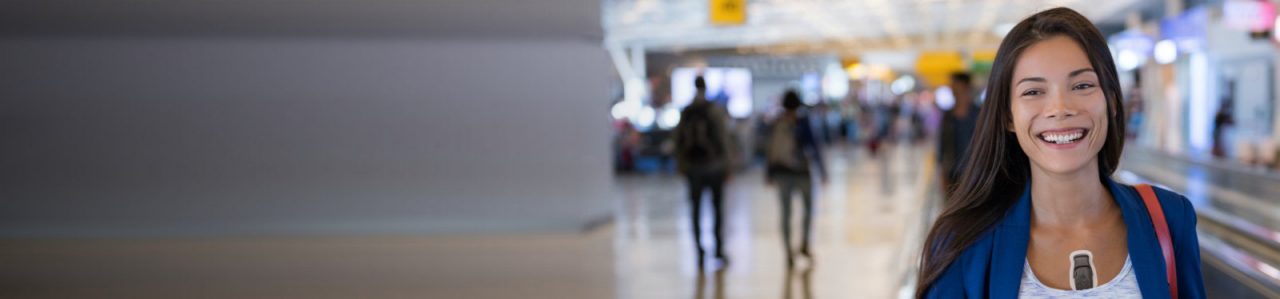 Woman confidently smiling in airport, wearing BodyGuardian Remote Cardiac Monitor.